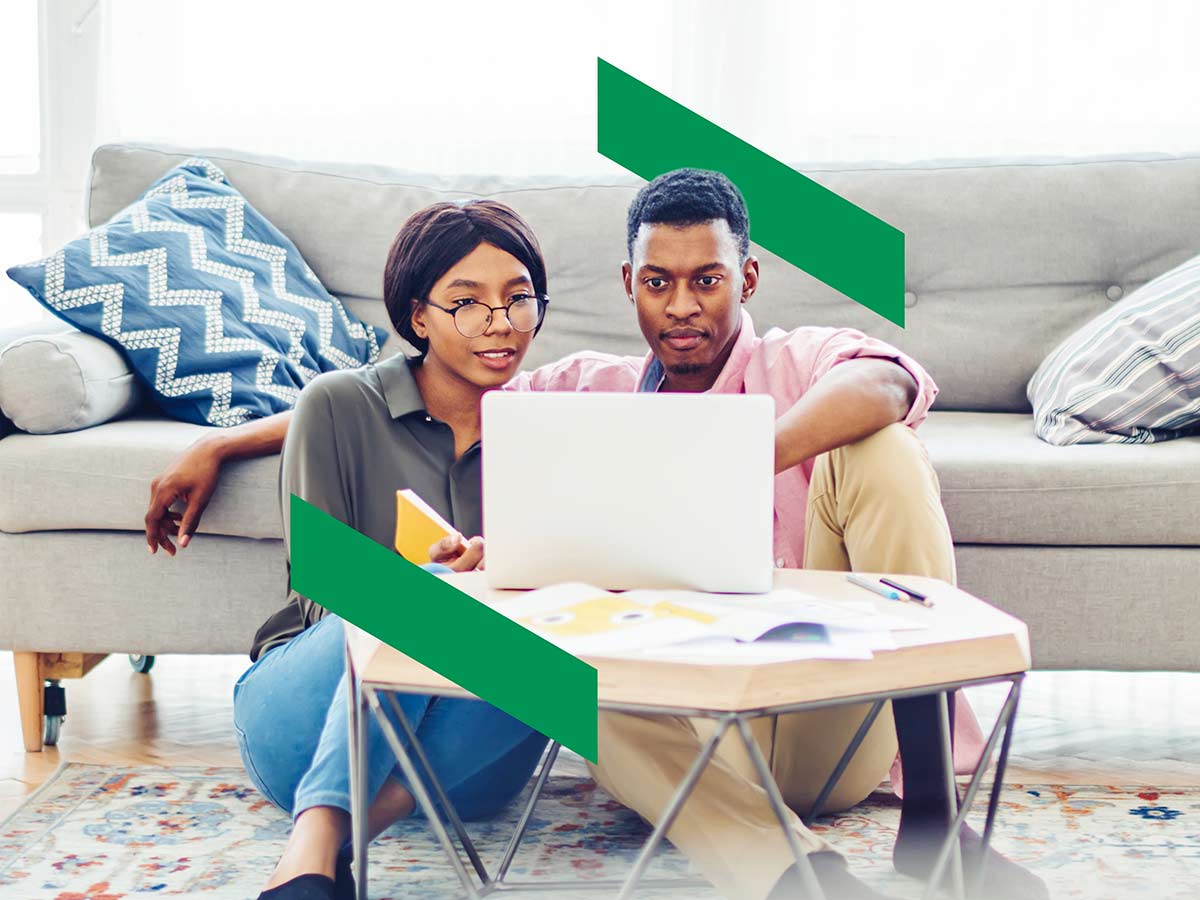 Couple sitting on the living room floor, leaning against a couch and looking at a computer screen.