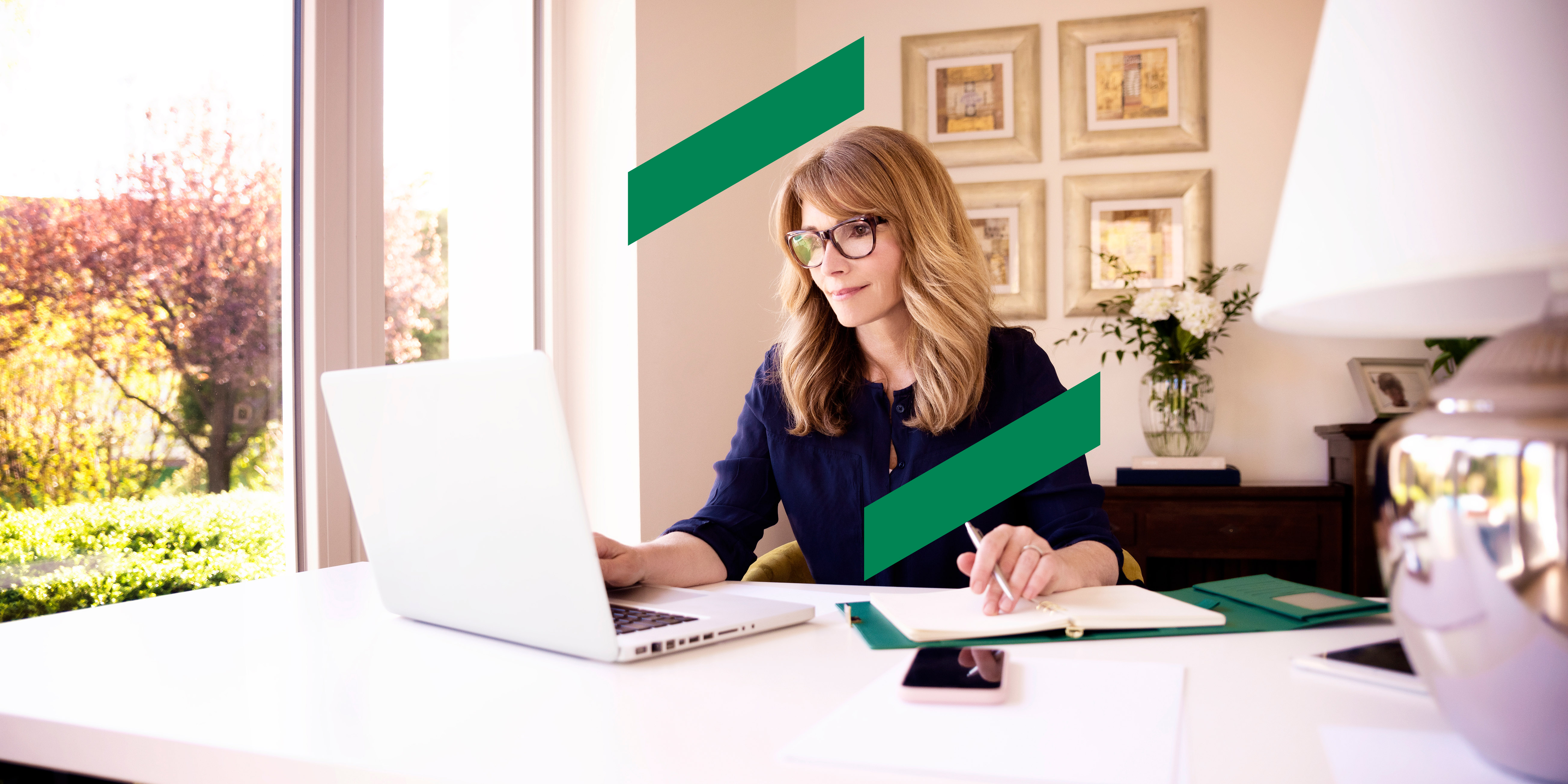 A woman wearing glasses is smiling as she works from a laptop and writes in a notebook.