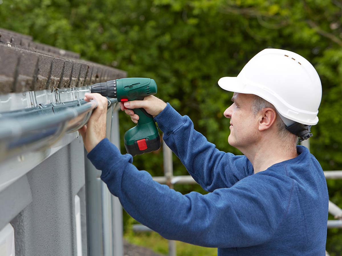 Man holding a power drill attaching gutters on a house.